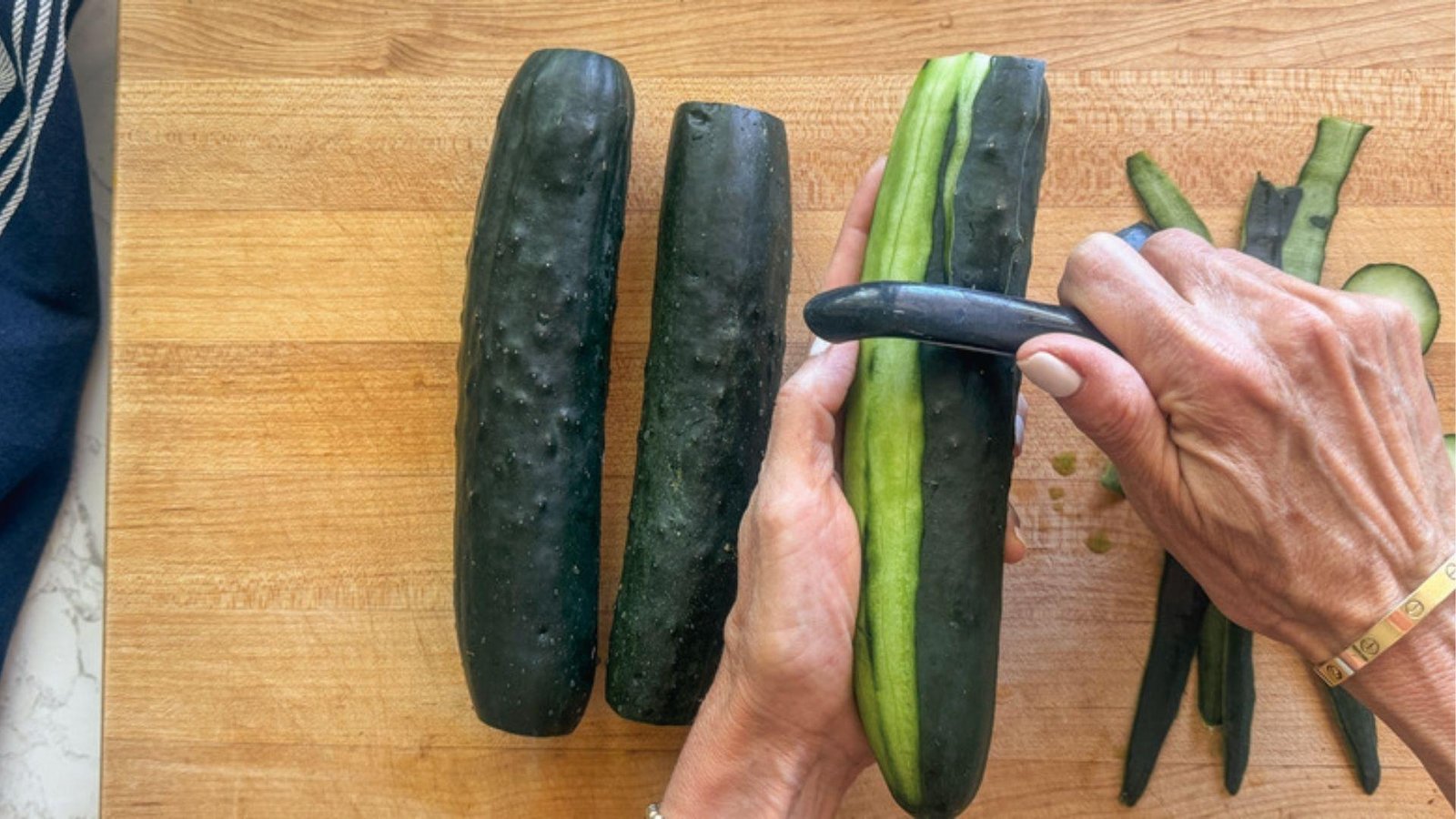 Hands peeling fresh cucumbers on wooden board with peels aside for cucumber mint agua fresca prep