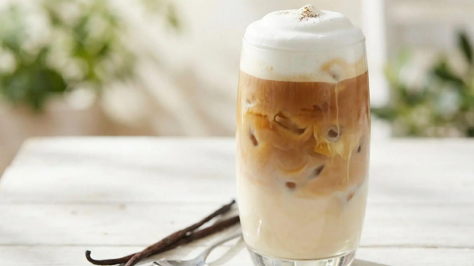 A tall iced vanilla latte with thick foam and a cinnamon dusting, with vanilla bean pods on a white surface.