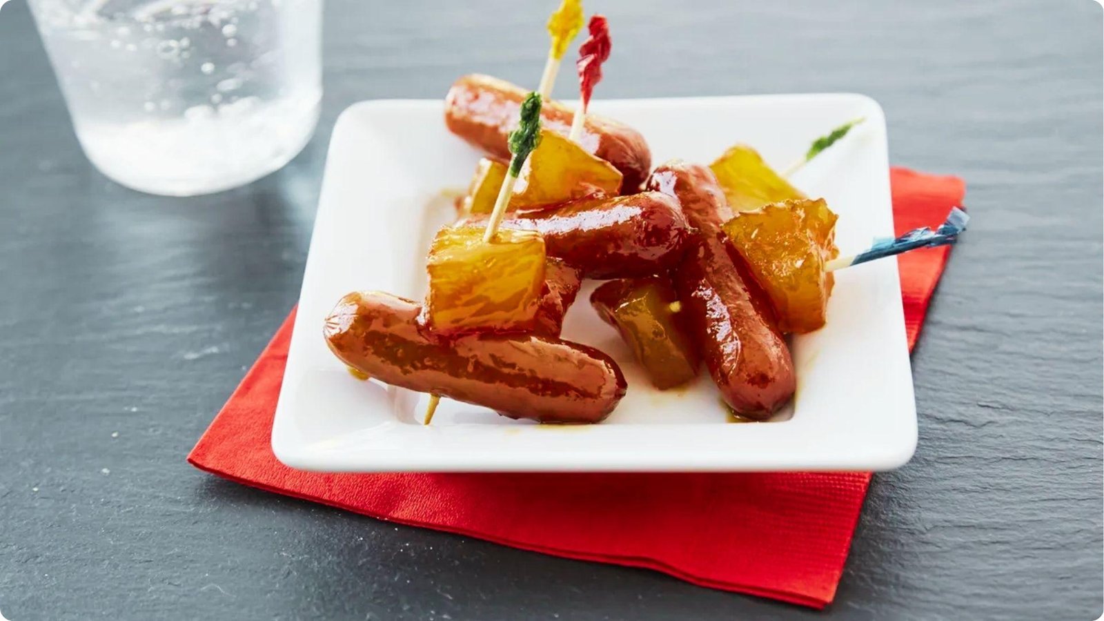 Glazed cocktail sausages and caramelized pineapple chunks on colorful toothpicks served on a white plate with red napkin.