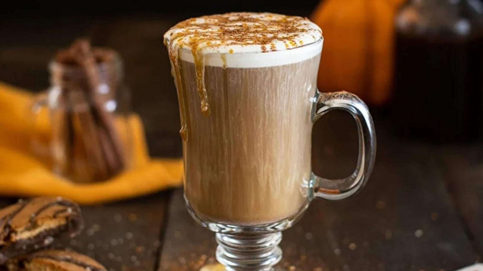 A tall glass mug of pumpkin spice latte with foam and caramel drips, surrounded by cinnamon sticks and a pumpkin.