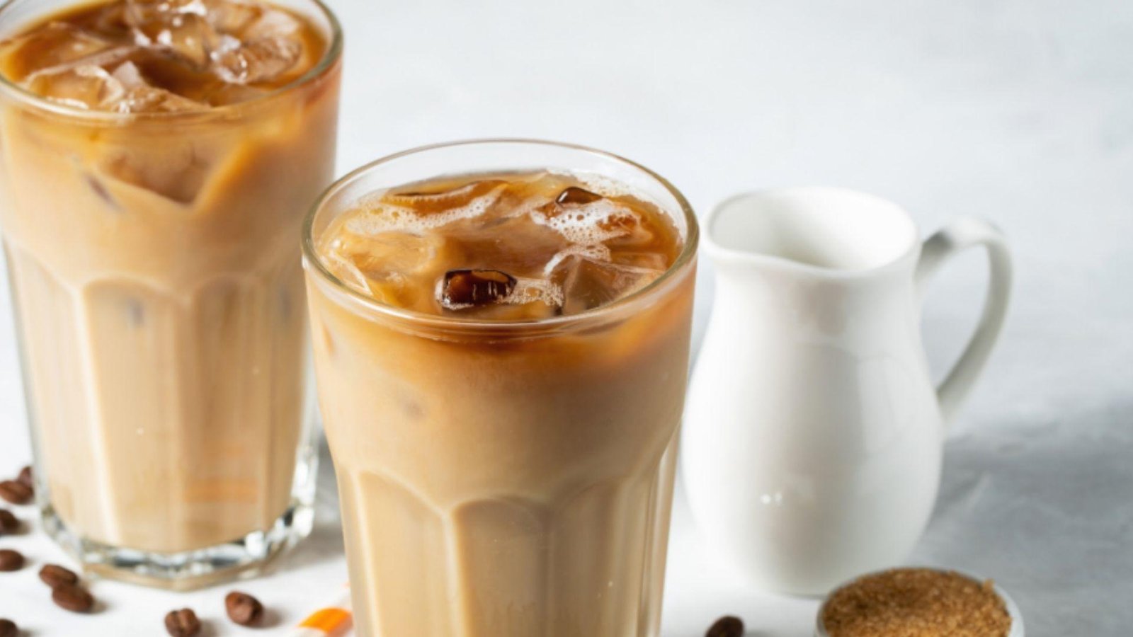 Two tall iced coffee glasses with swirling milk and ice cubes beside a white cream pitcher on a light surface.
