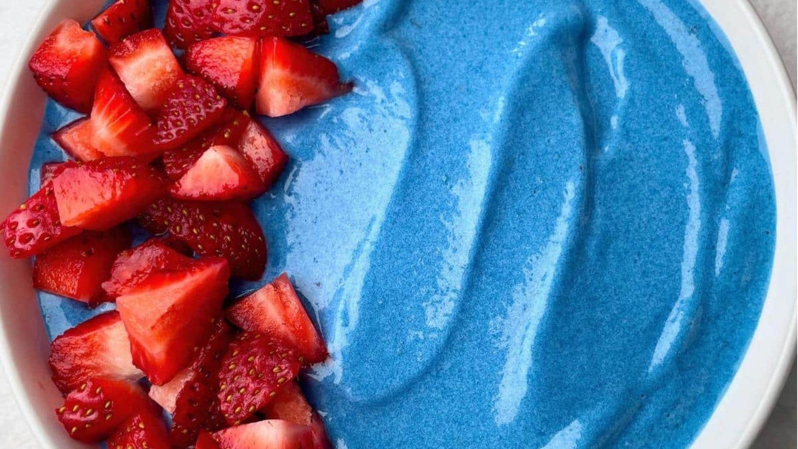Close-up of a vibrant blue spirulina smoothie bowl in a white bowl topped with fresh chopped red strawberries on one side.