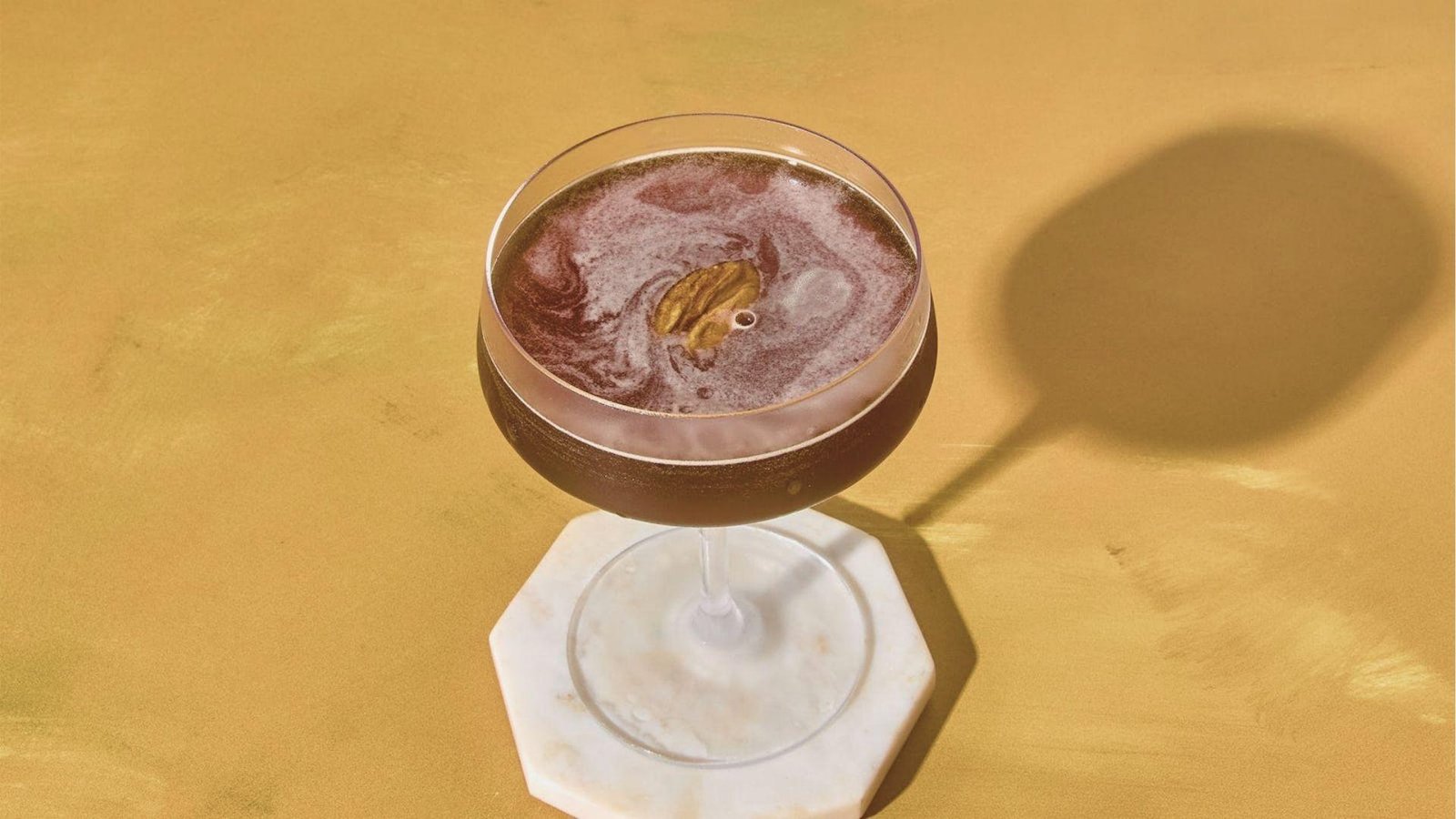 Mezcal Espresso Martini in a coupe glass with swirled foam and a cardamom pod garnish, set on a white marble coaster.