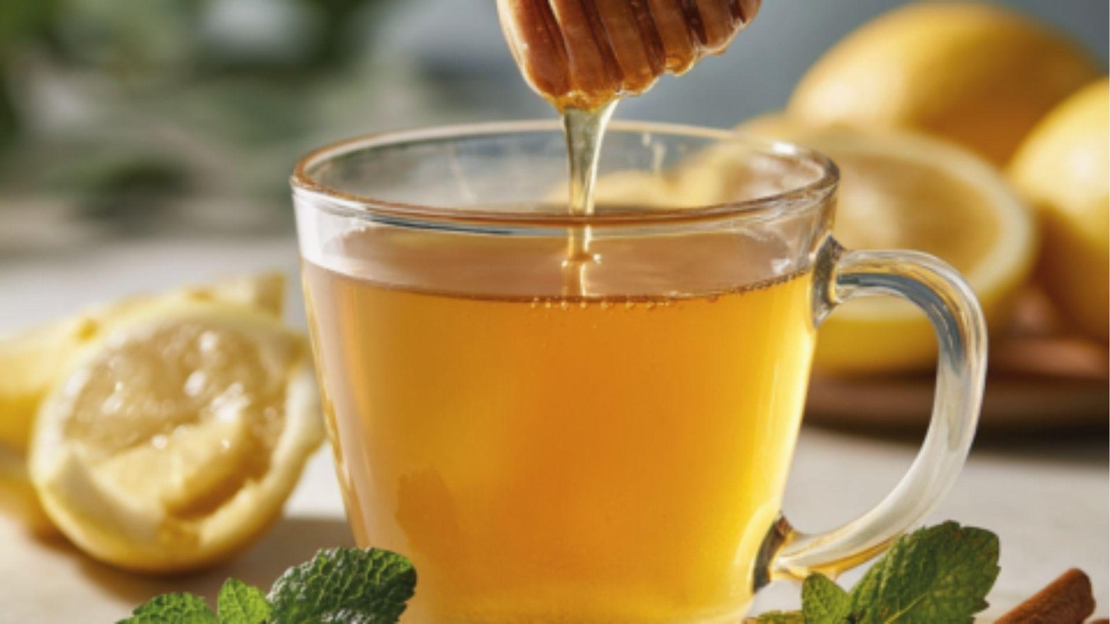 Honey drips from the dipper into a glass mug of warm lemon water with mint and cinnamon sticks.