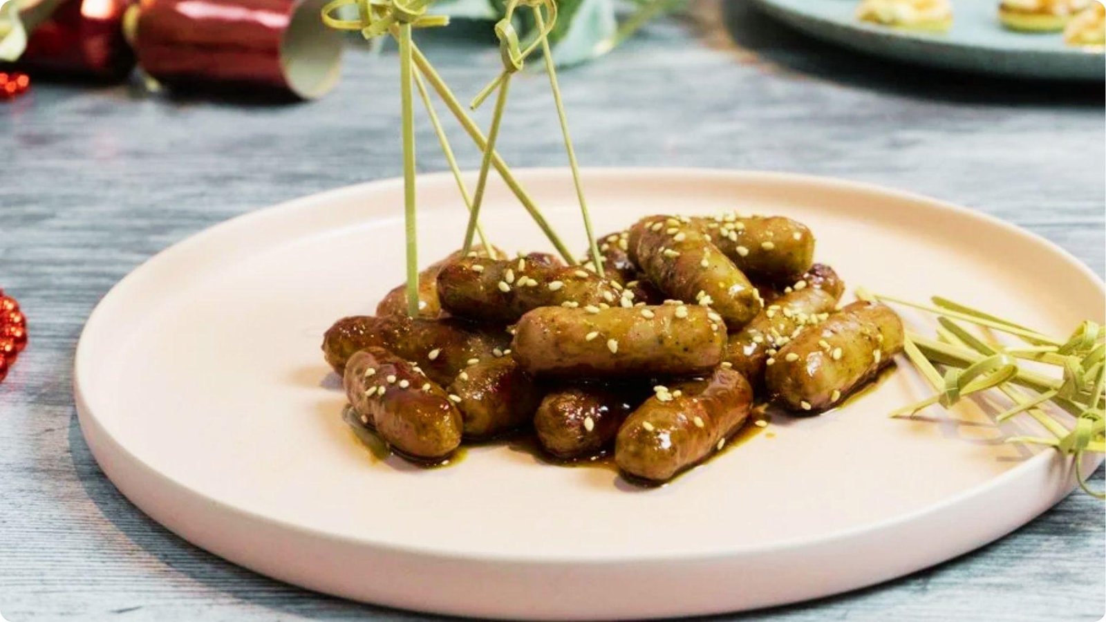 Sesame-sprinkled glazed cocktail sausages on decorative picks piled on a round plate with Christmas crackers in background.