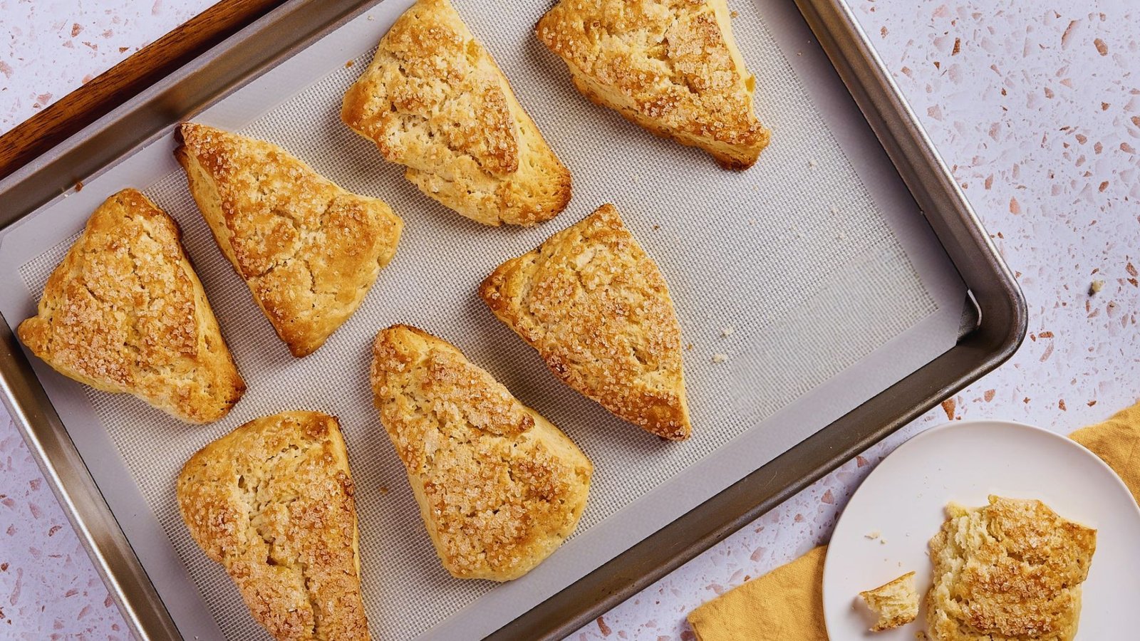 Eight golden ginger scones with coarse sugar on baking sheet, one broken on a plate beside.