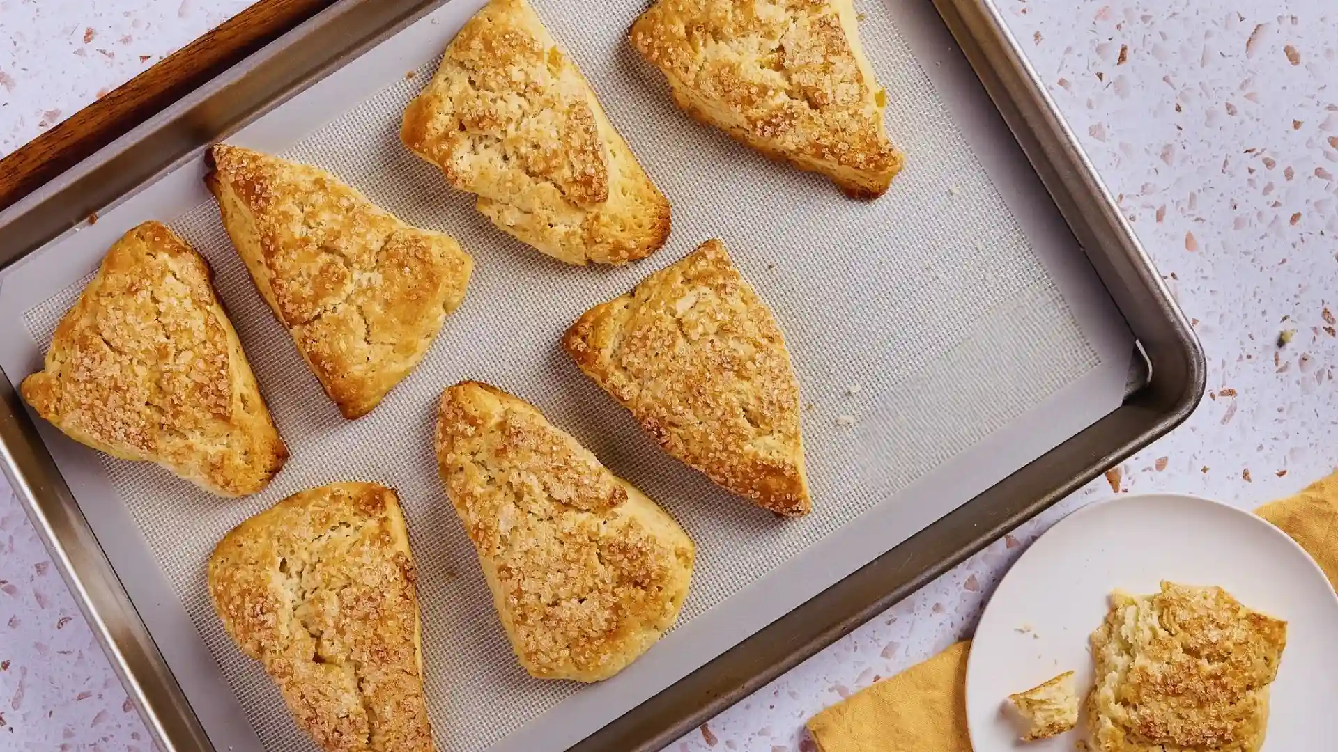 Eight golden ginger scones with coarse sugar on baking sheet, one broken on a plate beside.