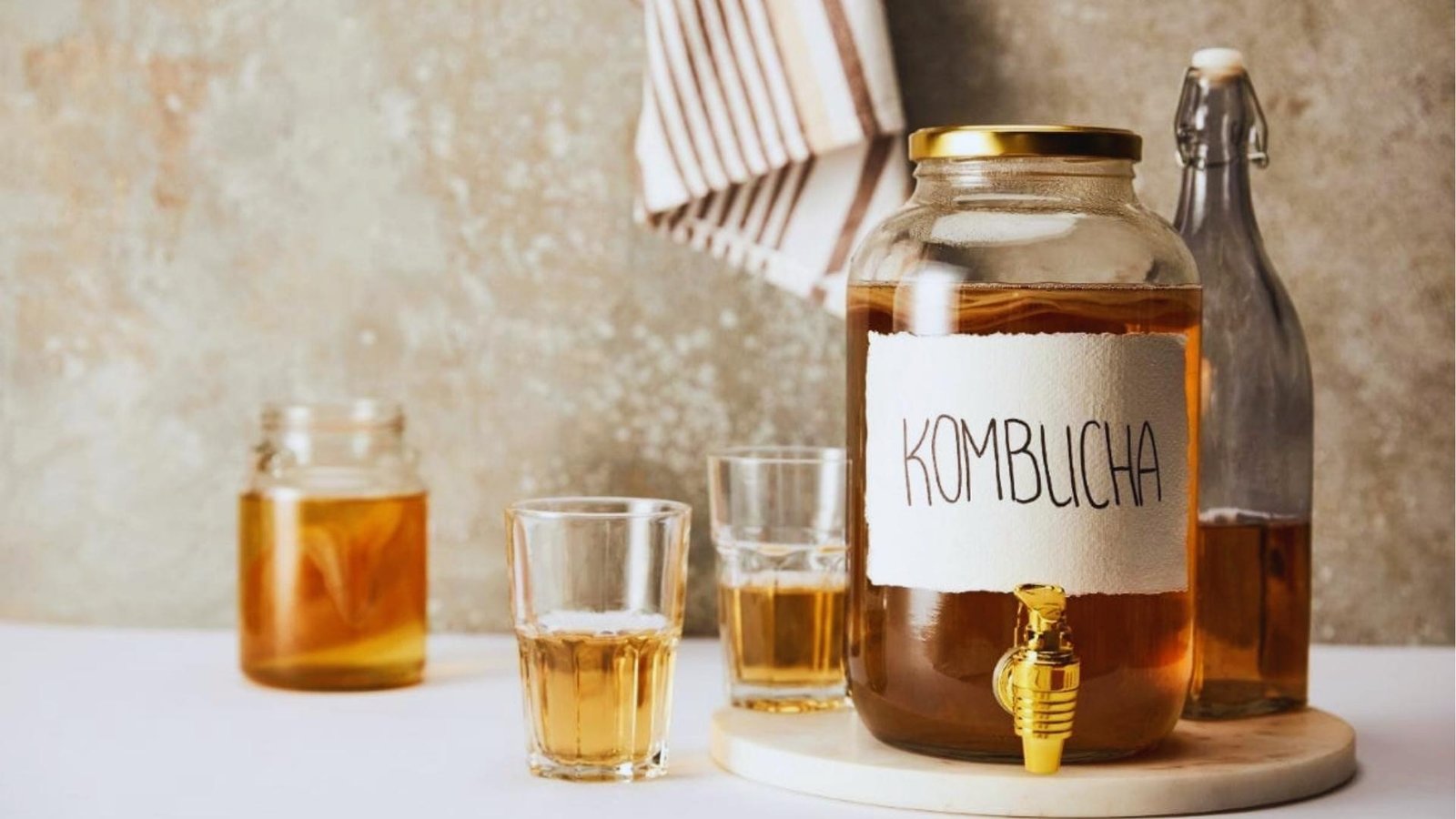 A large glass kombucha dispenser with a gold spigot labeled "KOMBUCHA" sits beside filled glasses and a swing-top bottle on a marble board.