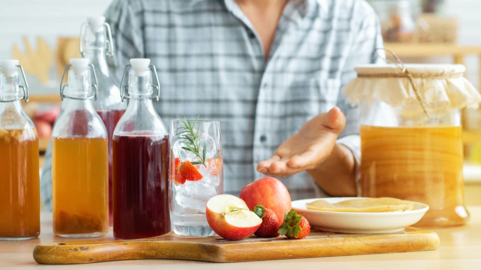 Person presenting homemade kombucha bottles, fresh fruit, and SCOBY jar on kitchen counter during brewing process.