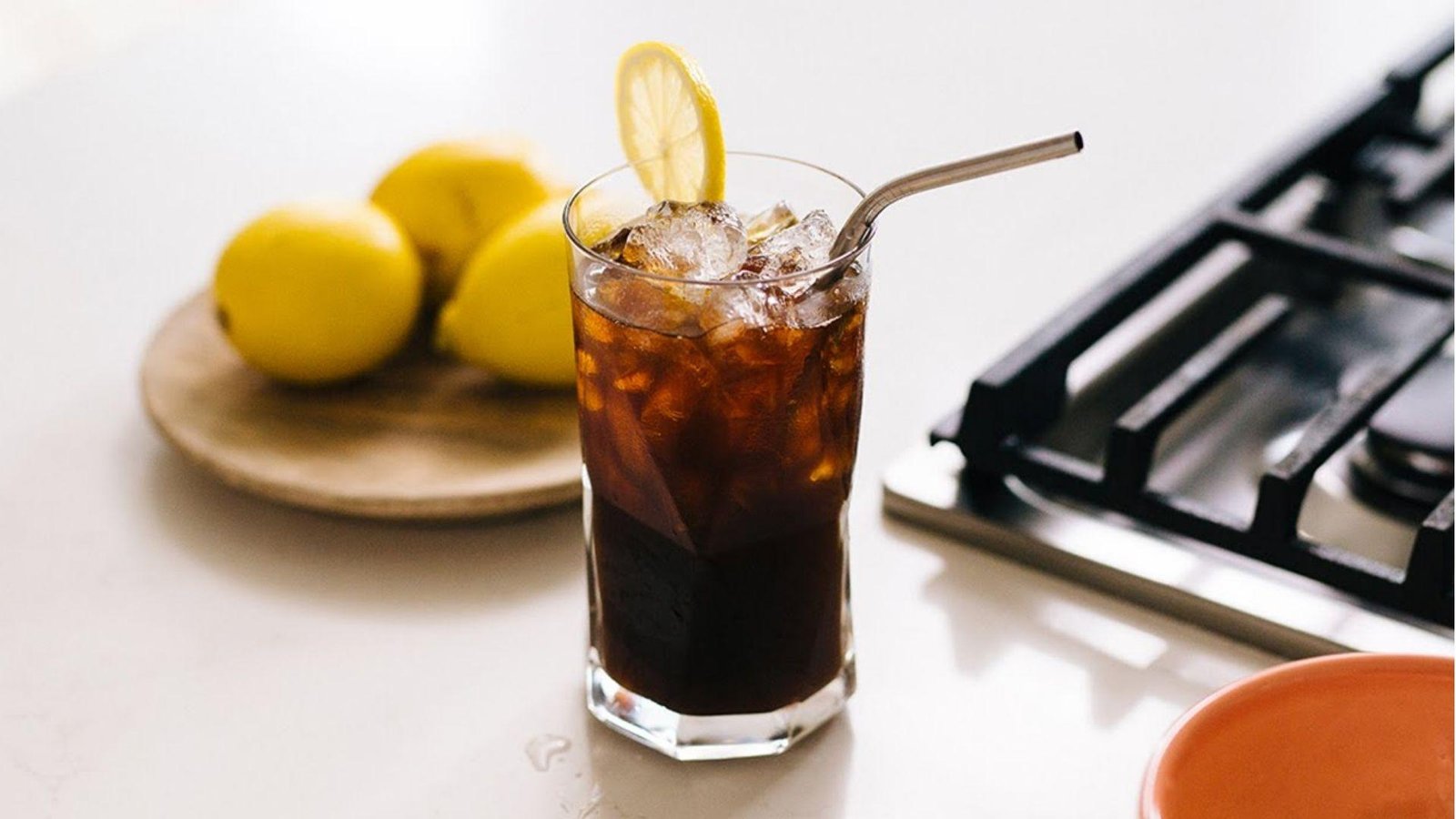A tall glass of iced black coffee garnished with a lemon slice and metal straw, with lemons in the background.
