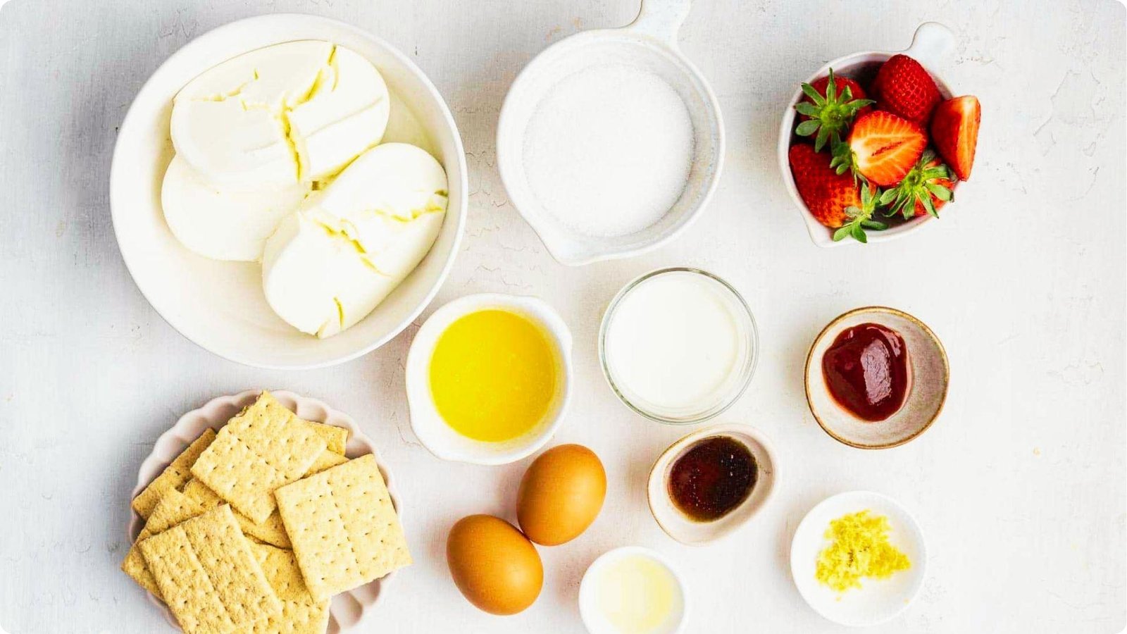 Top-down view of 6-inch cheesecake ingredients arranged in bowls: cream cheese, sugar, fresh strawberries, eggs, and graham crackers.