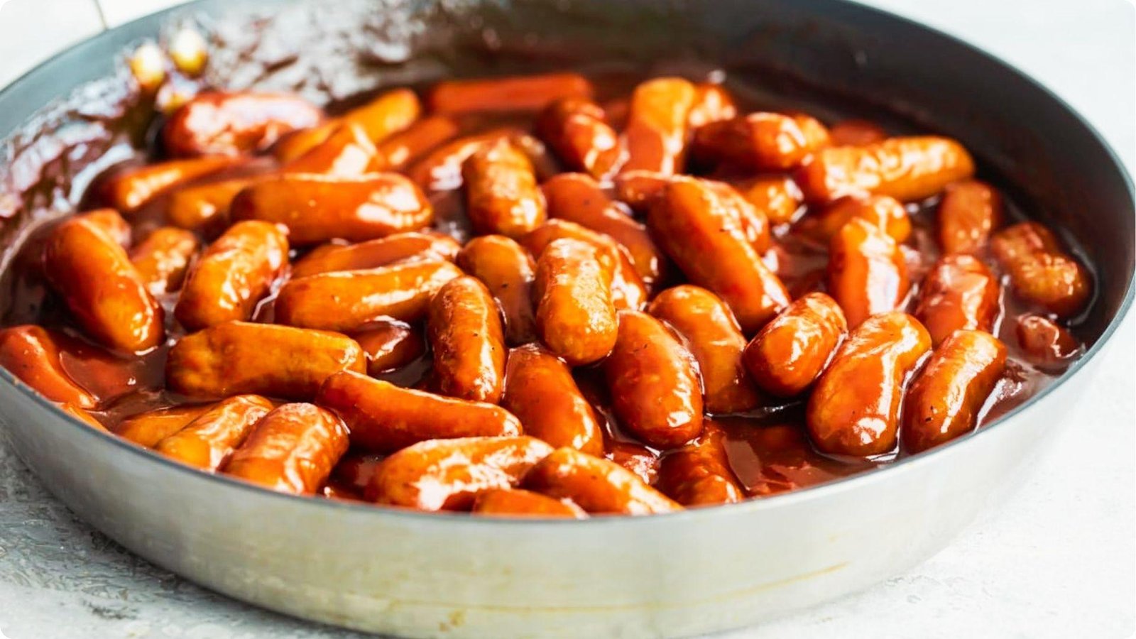 Cocktail sausages simmering in a thick, glossy BBQ sauce in a large skillet, ready to serve.