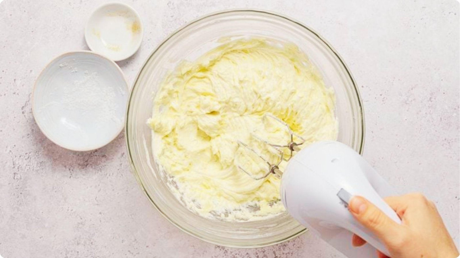 A hand mixer blends cream cheese in a glass bowl until smooth and fluffy for the 6-inch cheesecake filling.