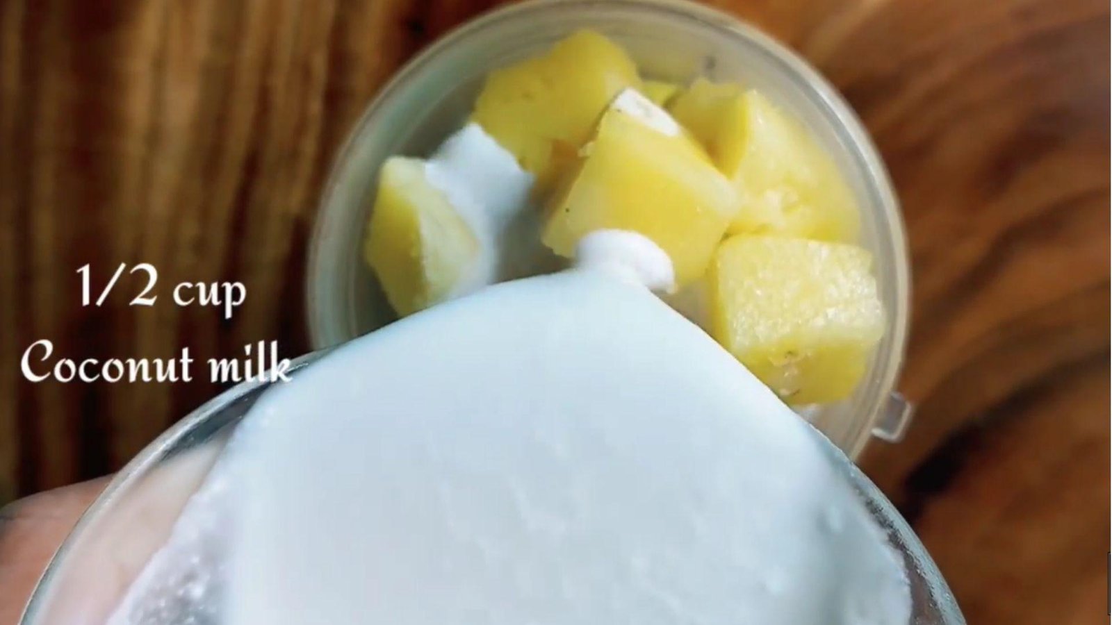 A hand pouring half a cup of coconut milk into a blender cup containing frozen pineapple chunks on a wooden surface.