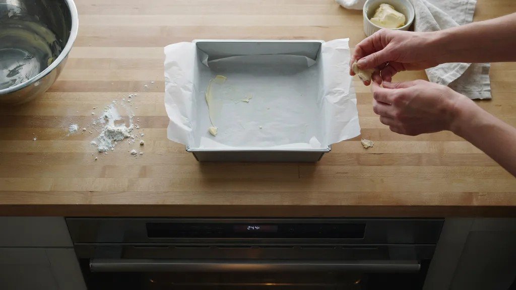 Hands greasing a parchment-lined square baking pan on a wooden countertop above an oven preheated to 244 degrees.