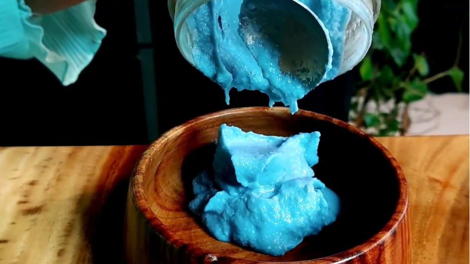 A thick vibrant blue spirulina smoothie being poured out of a blender cup into a round wooden bowl.