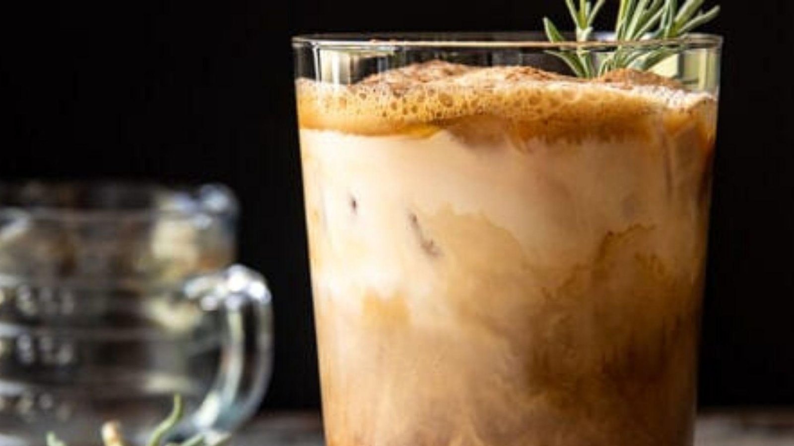 A rocks glass of dark iced coffee garnished with an orange wedge, with coffee foam on top, on a grey surface.