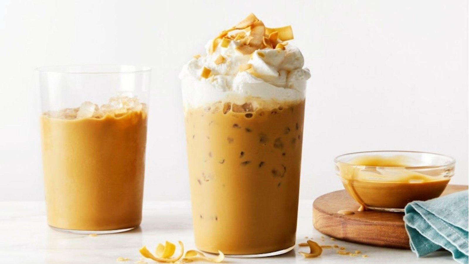 A tall iced caramel coffee topped with whipped cream and toasted coconut flakes, beside a small bowl of caramel sauce.