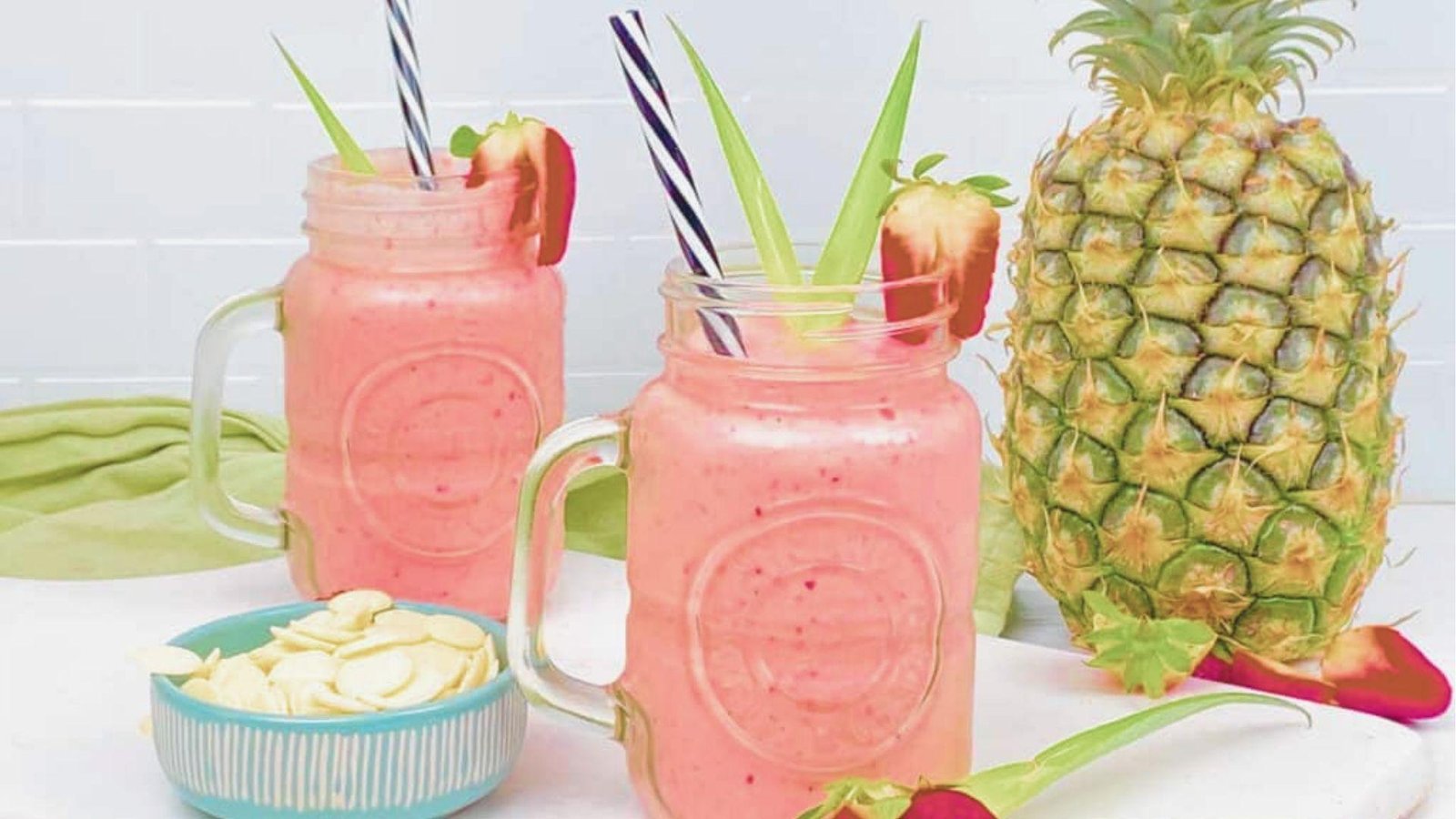 Two mason jar glasses of pink Bahama Mama Tropical Smoothie garnished with strawberry slices and striped straws.
