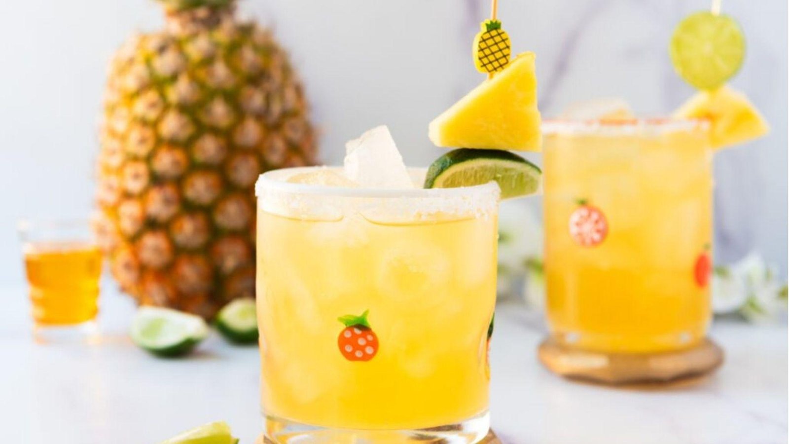 Two salted-rim rocks glasses of golden pineapple cocktail garnished with lime and pineapple wedges beside a whole pineapple.