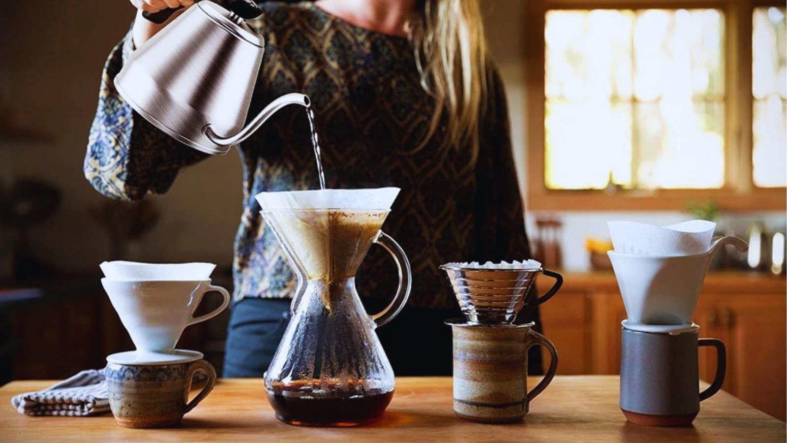 Person pours hot water from gooseneck kettle into Chemex pour-over brewer with filter, surrounded and Kalita drippers.