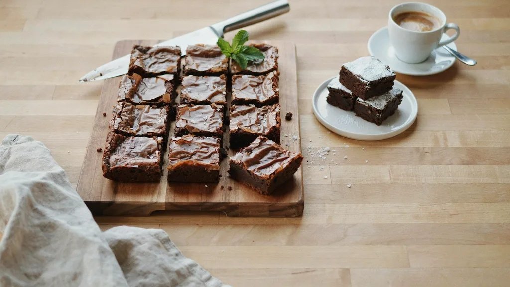 Fudgy brownies sliced on a wooden board with mint garnish, served alongside a white espresso cup on a counter.