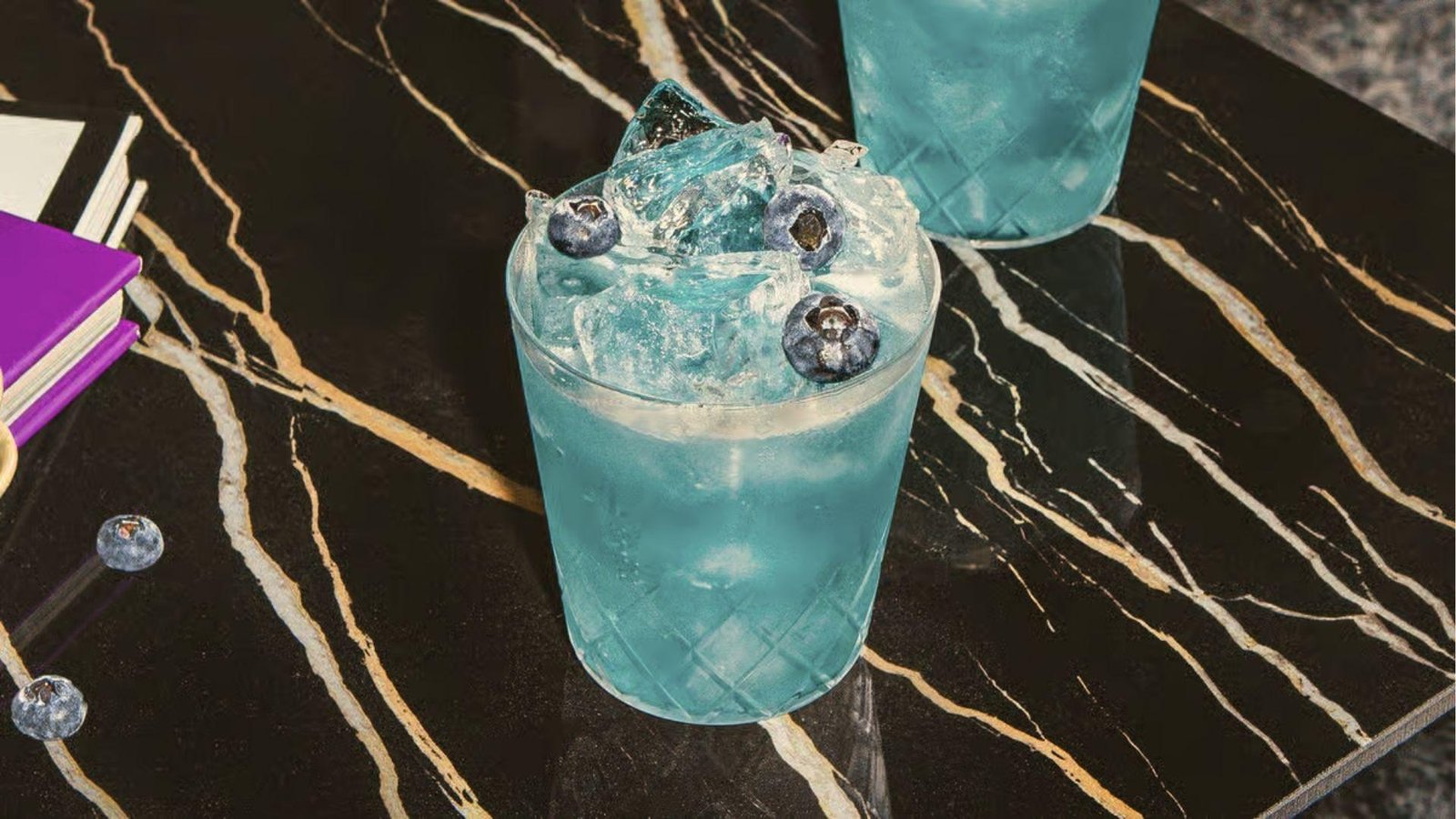 Blue salt trick cocktail with blueberries and ice in crystal glass on black marble with gold veins