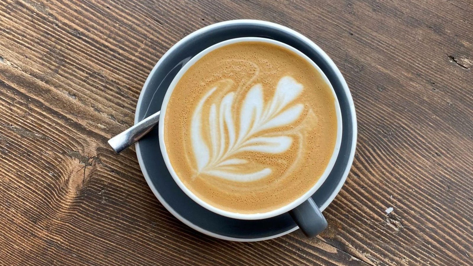 An overhead view of a latte with intricate rosette latte art in a grey ceramic cup and saucer with a silver spoon, placed on a rustic wooden table.
