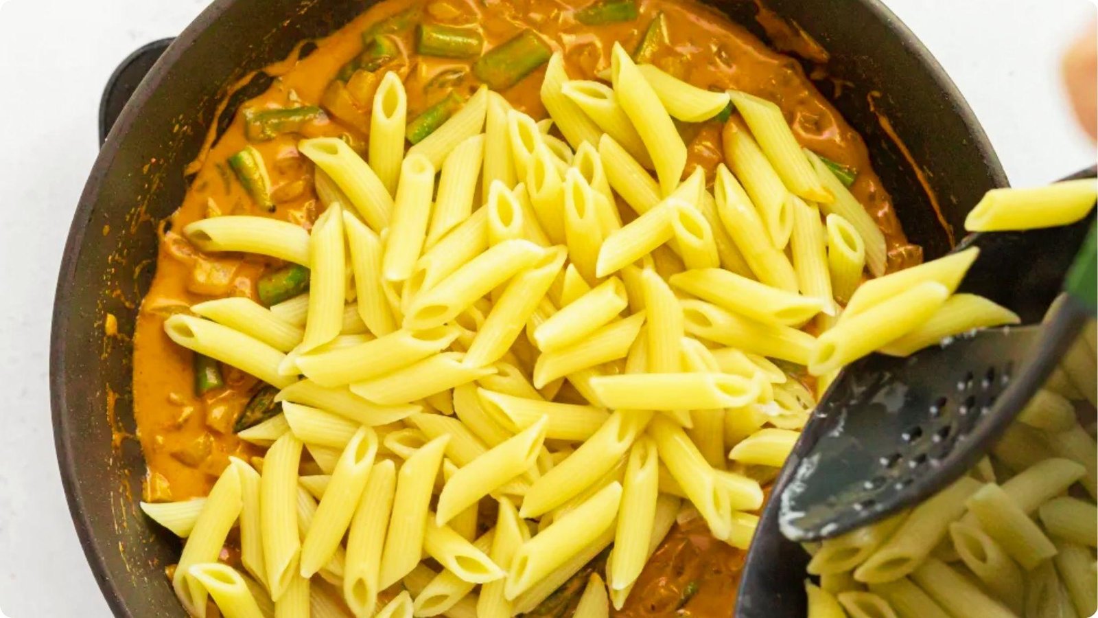 Cooked pasta being added to a skillet of creamy chipotle sauce with asparagus and onions.