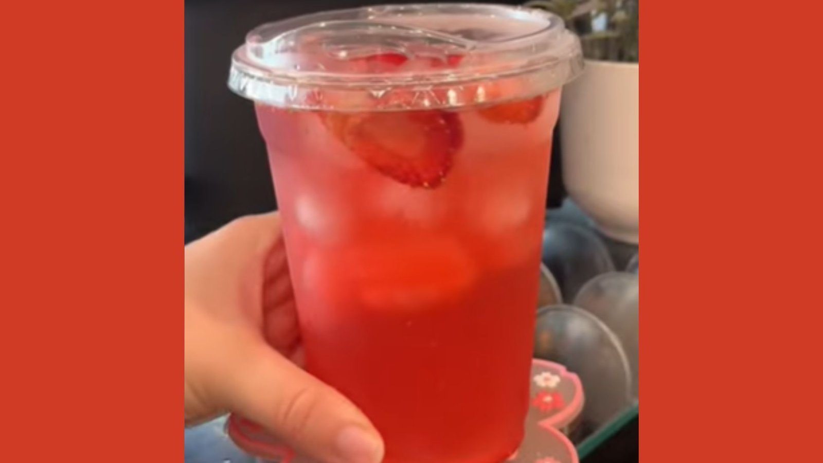 A hand holding a clear plastic cup of red strawberry iced drink with strawberry slices visible through the cup.