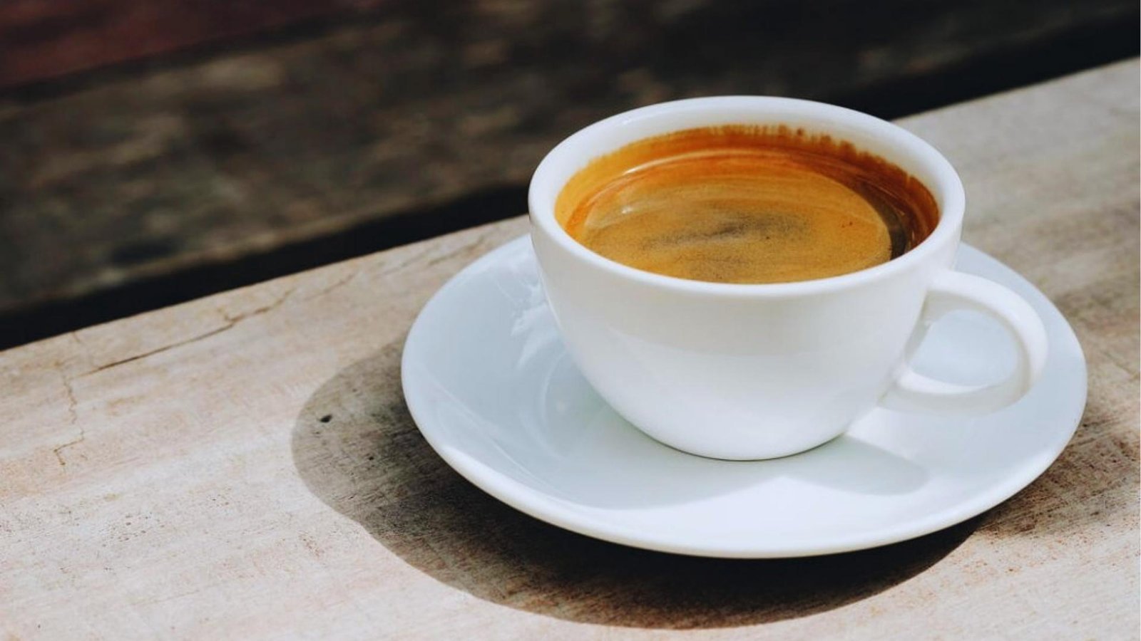 White ceramic cup of Americano with golden crema sits on a saucer in natural light on a weathered wood table outdoors.