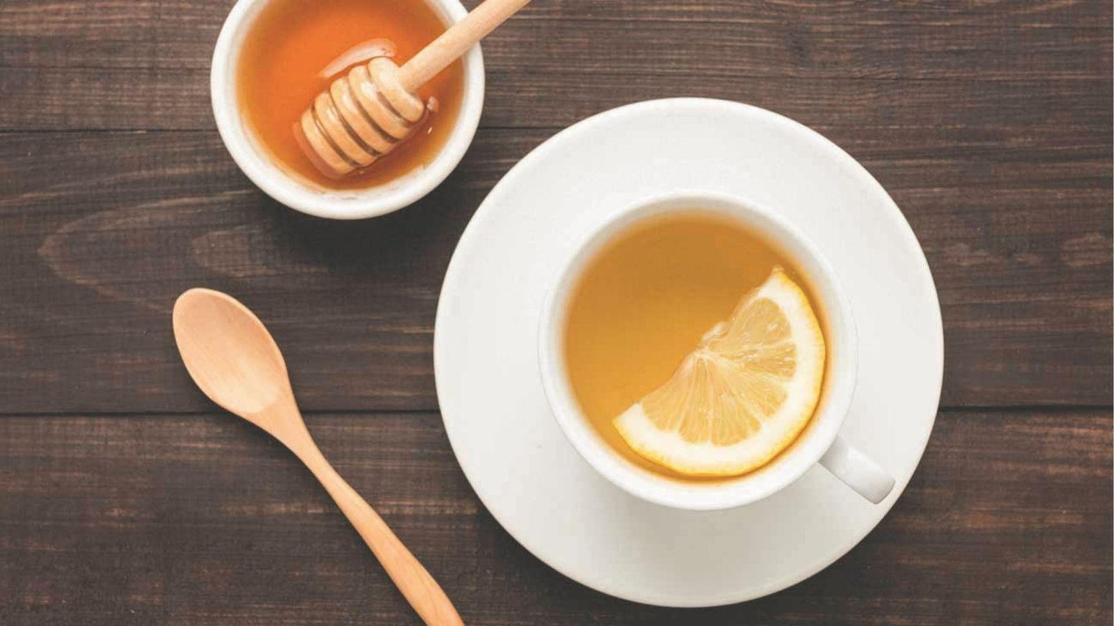 White teacup with lemon slice next to honey bowl and spoon on dark wood for honey trick.