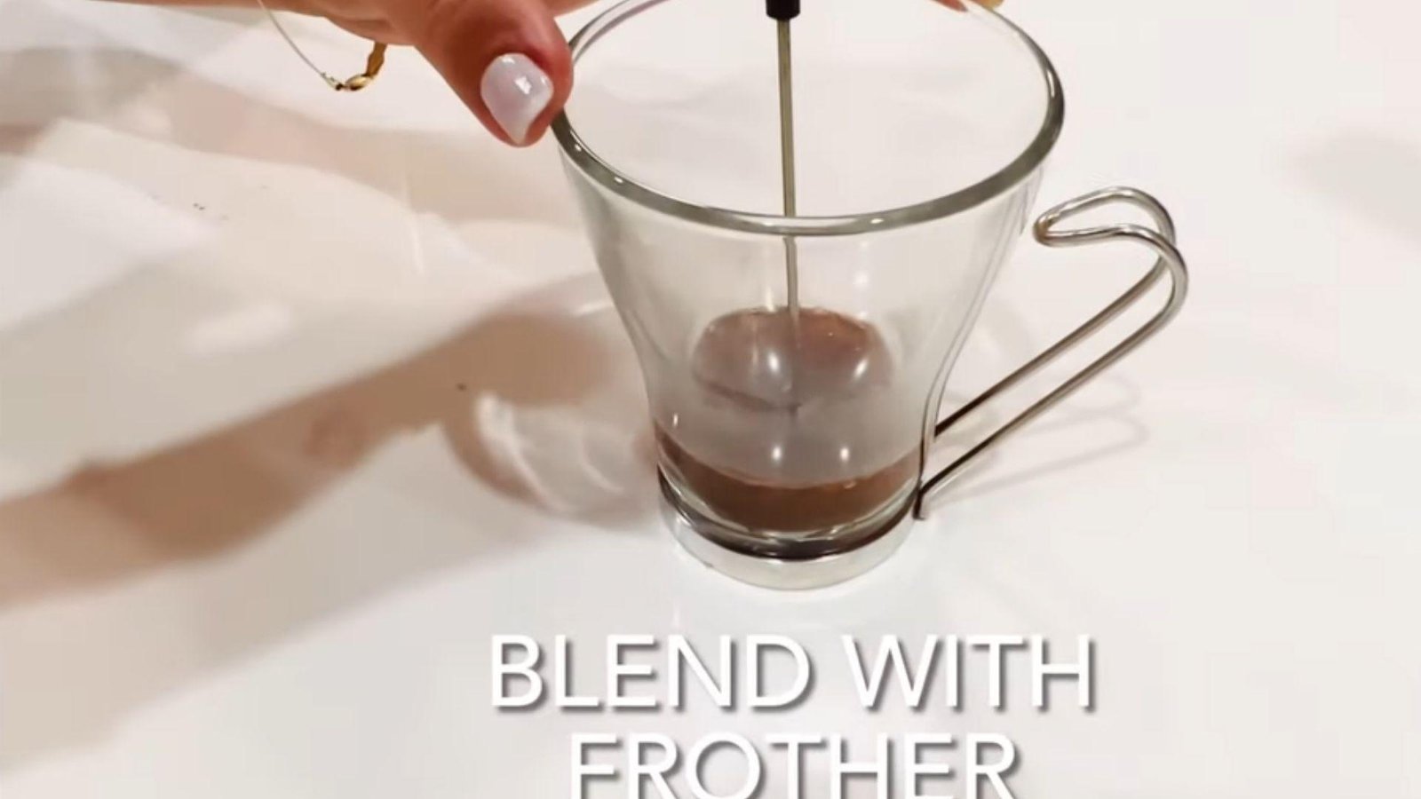 A hand using a milk frother to blend ingredients in a clear glass mug with text reading "Blend with frother" below.