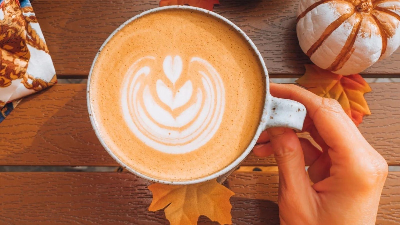 Overhead view of a hand holding a white mug of pumpkin spice latte with tulip latte art, surrounded by autumn leaves.