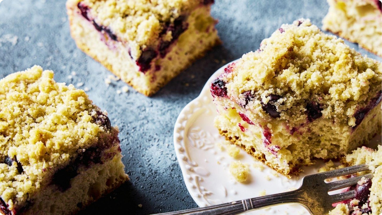 Squares of German berry coffee cake with blueberry filling and a generous streusel topping on a dark slate surface.