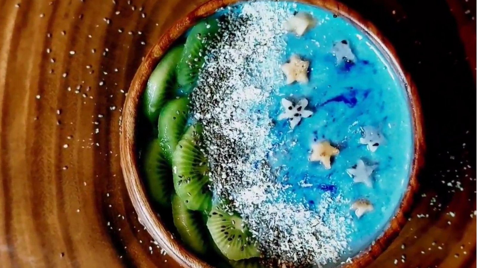 Overhead view of a finished blue spirulina smoothie bowl topped with kiwi slices, chia seeds, shredded coconut, and star-shaped banana pieces.