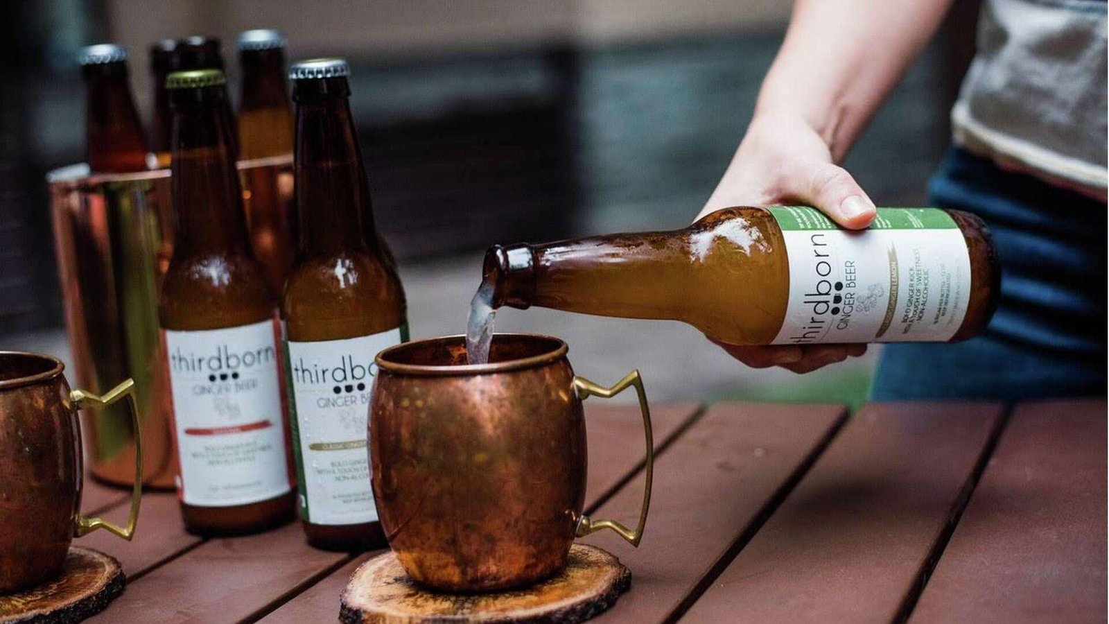 A hand pouring Thirdborn Ginger Beer into a copper Moscow mule mug on a wooden table with more bottles behind.