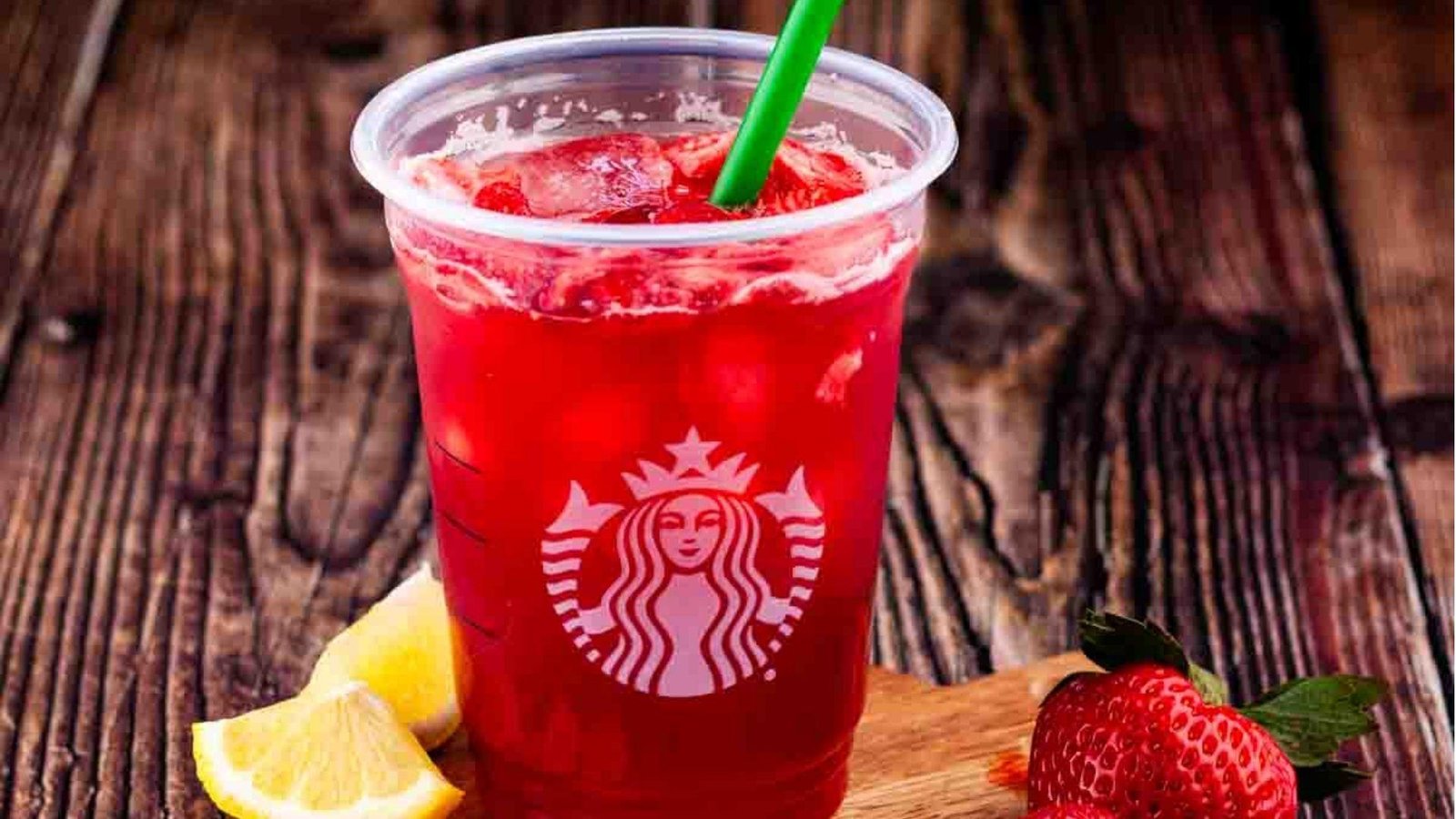 A Starbucks cup of bright red strawberry iced drink with a green straw, beside lemon slices and a fresh strawberry.