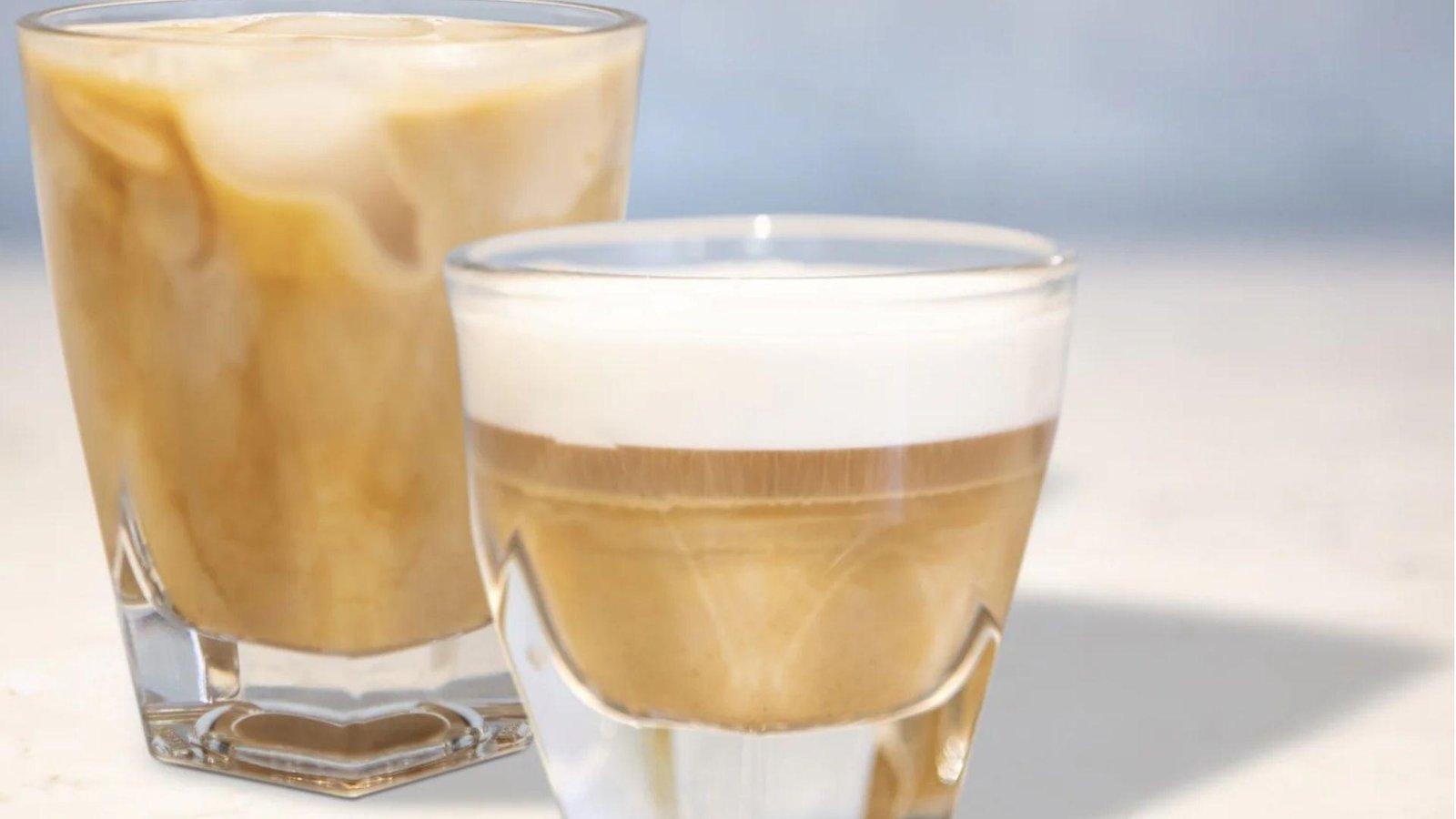 A small layered apple pie latte glass beside a larger iced coffee glass with swirling cream on a light blue surface.