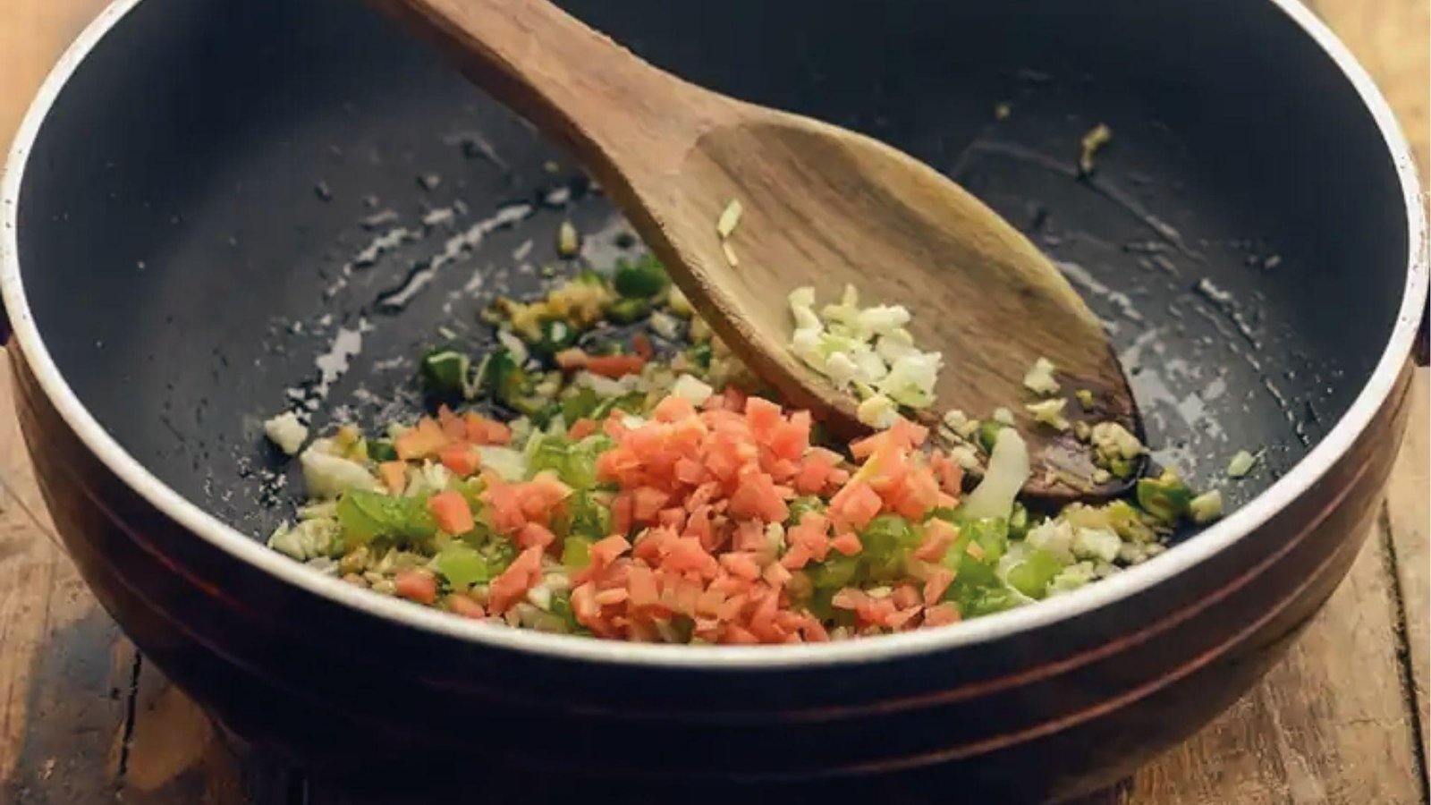 Sautéing diced carrots, celery, onions, and garlic in black pan with wooden spoon for dressing