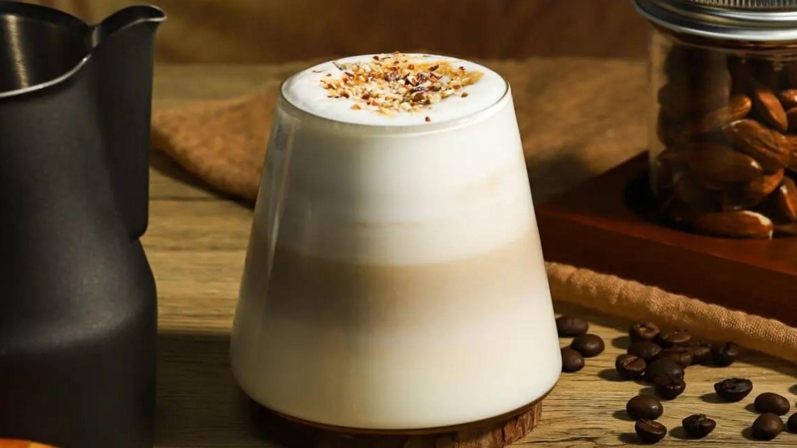 A layered white maple pecan latte topped with crushed mixed nuts in a tapered glass, surrounded by coffee beans and almonds.