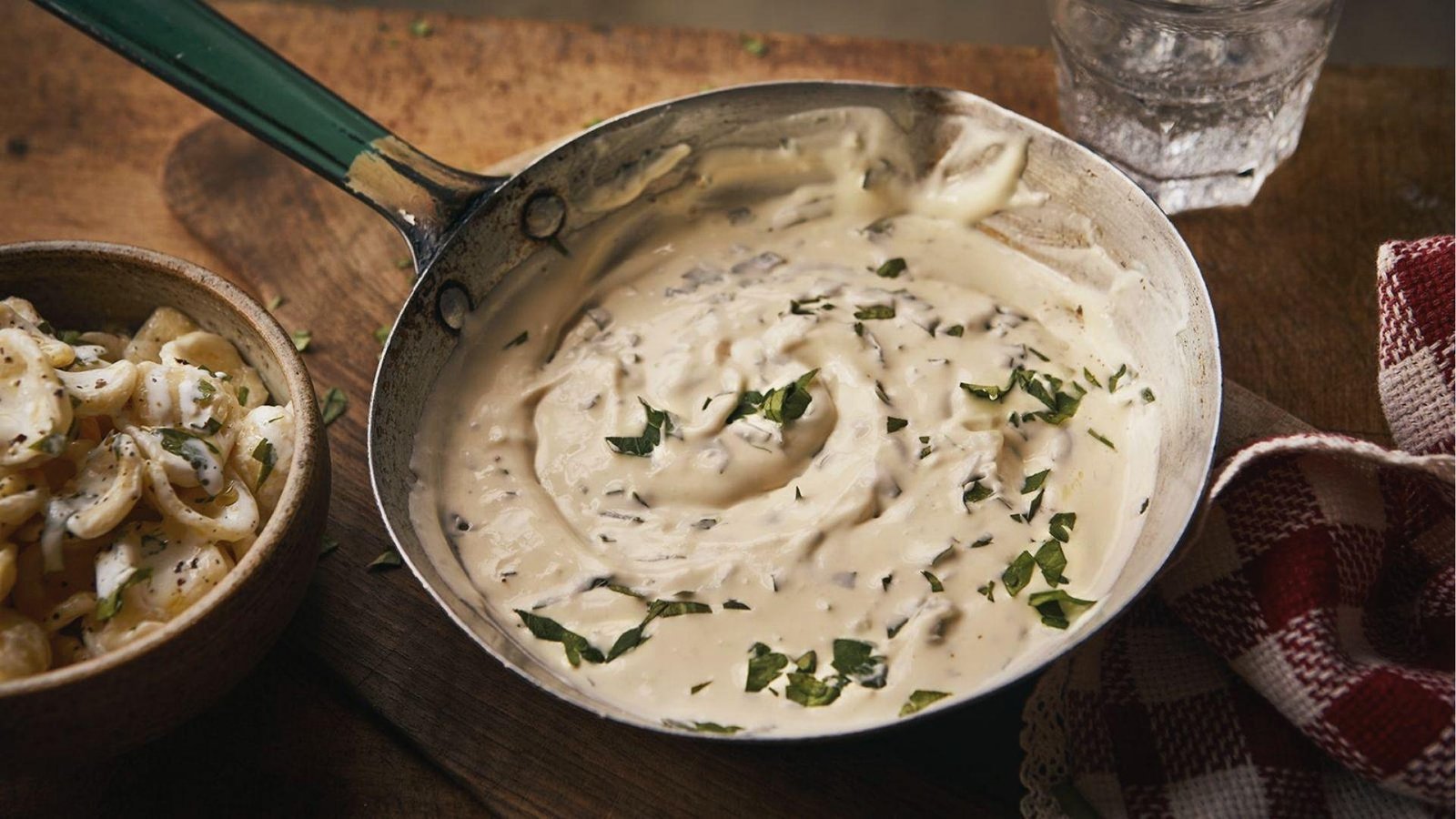 Thick herb cream sauce swirled in vintage pan beside bowl of creamy shell pasta on wood board