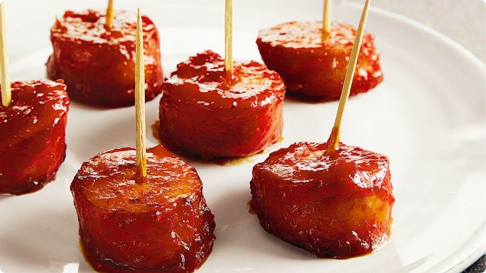Six bourbon wrapped bites glazed in sticky BBQ sauce, each speared with a toothpick, served on a white plate.