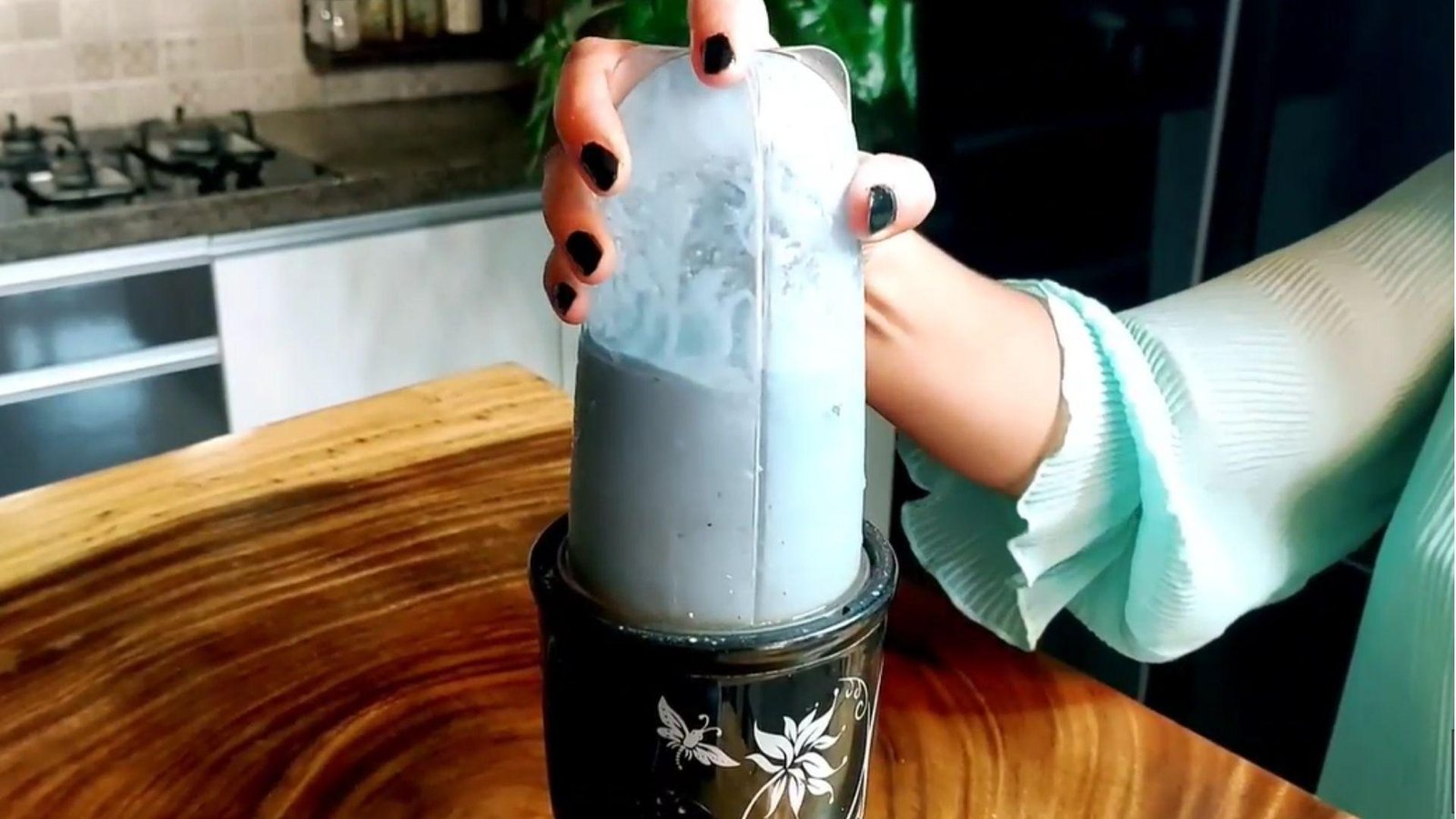 A woman's hands holding a blender cup filled with a pale blue spirulina smoothie mixture onto a blender base in a kitchen.