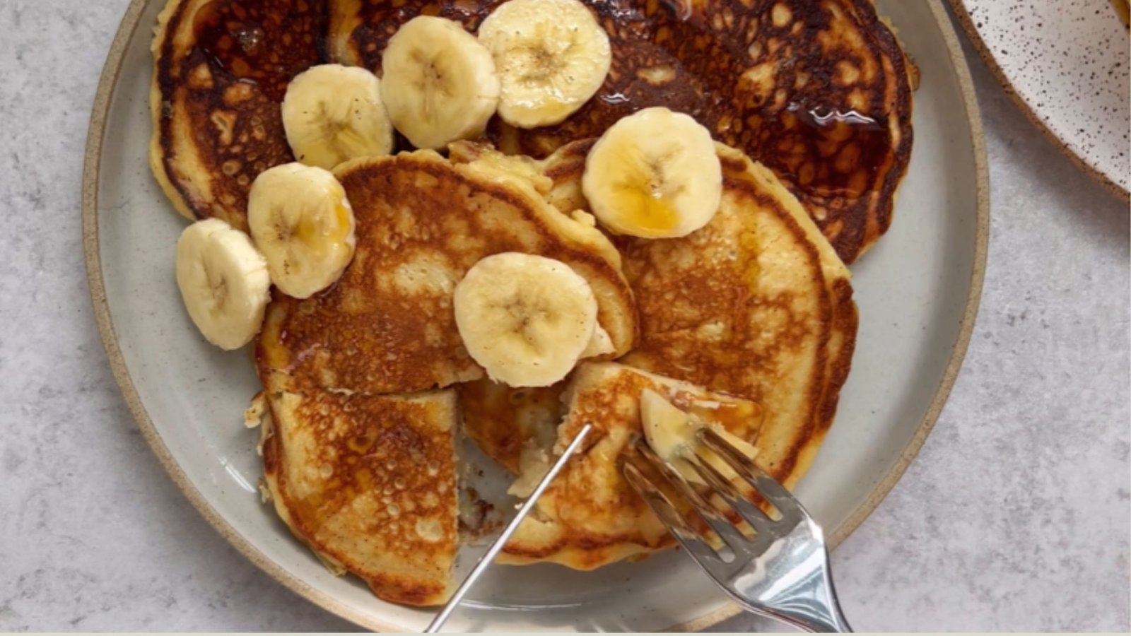 Overhead view of golden yogurt pancakes on ceramic plate topped with banana slices, maple syrup drizzle, and fork cutting into stack.
