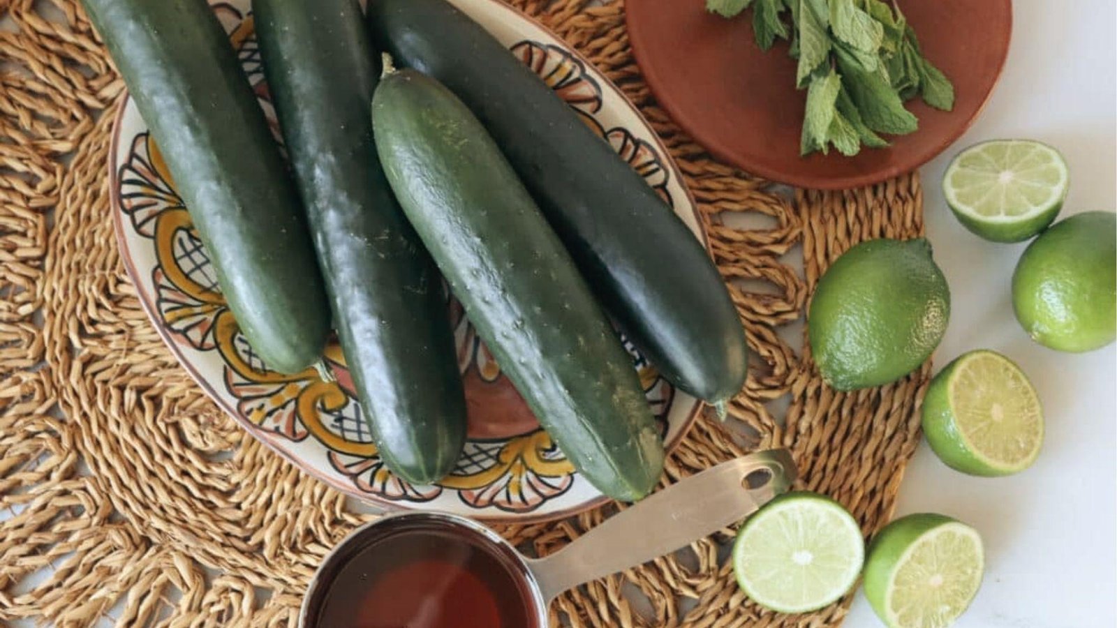 Fresh ingredients for cucumber mint agua fresca: cucumbers, limes, mint, and syrup on woven placemat