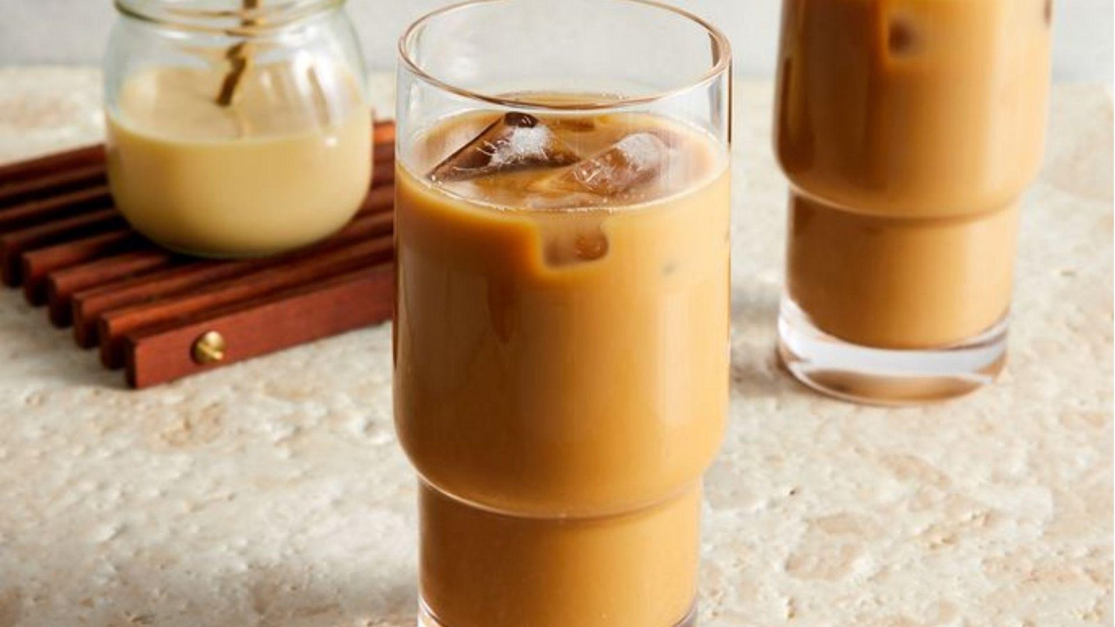 Two tall glasses of creamy iced coffee with ice cubes beside a jar of cream on a wooden trivet.