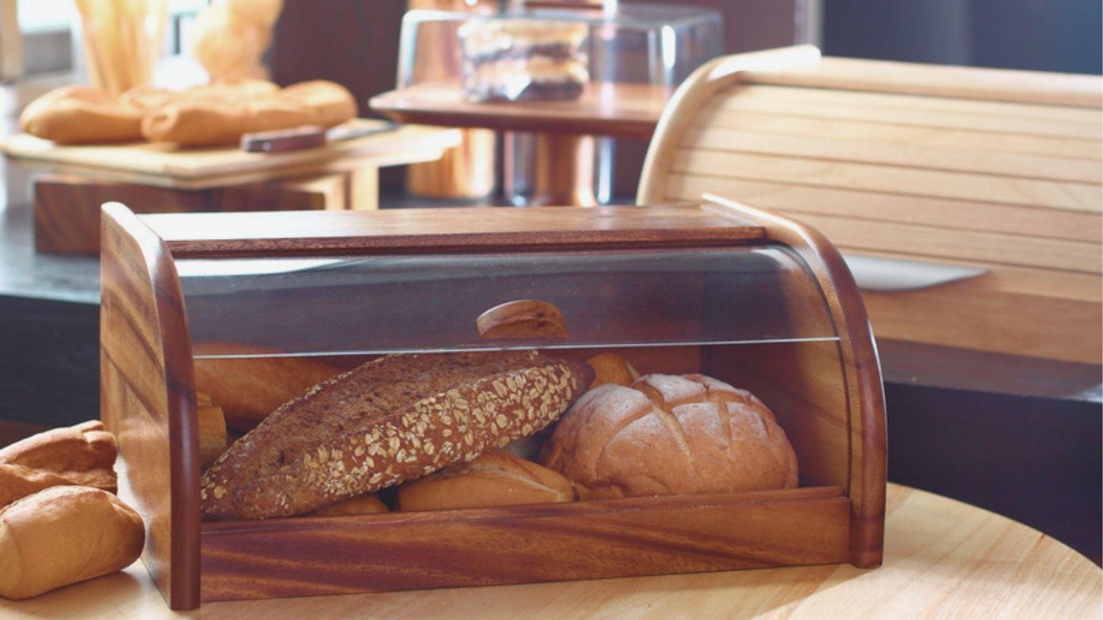 Wooden bread box with a clear lid storing assorted loaves, including a dark oat-topped coffee bread.