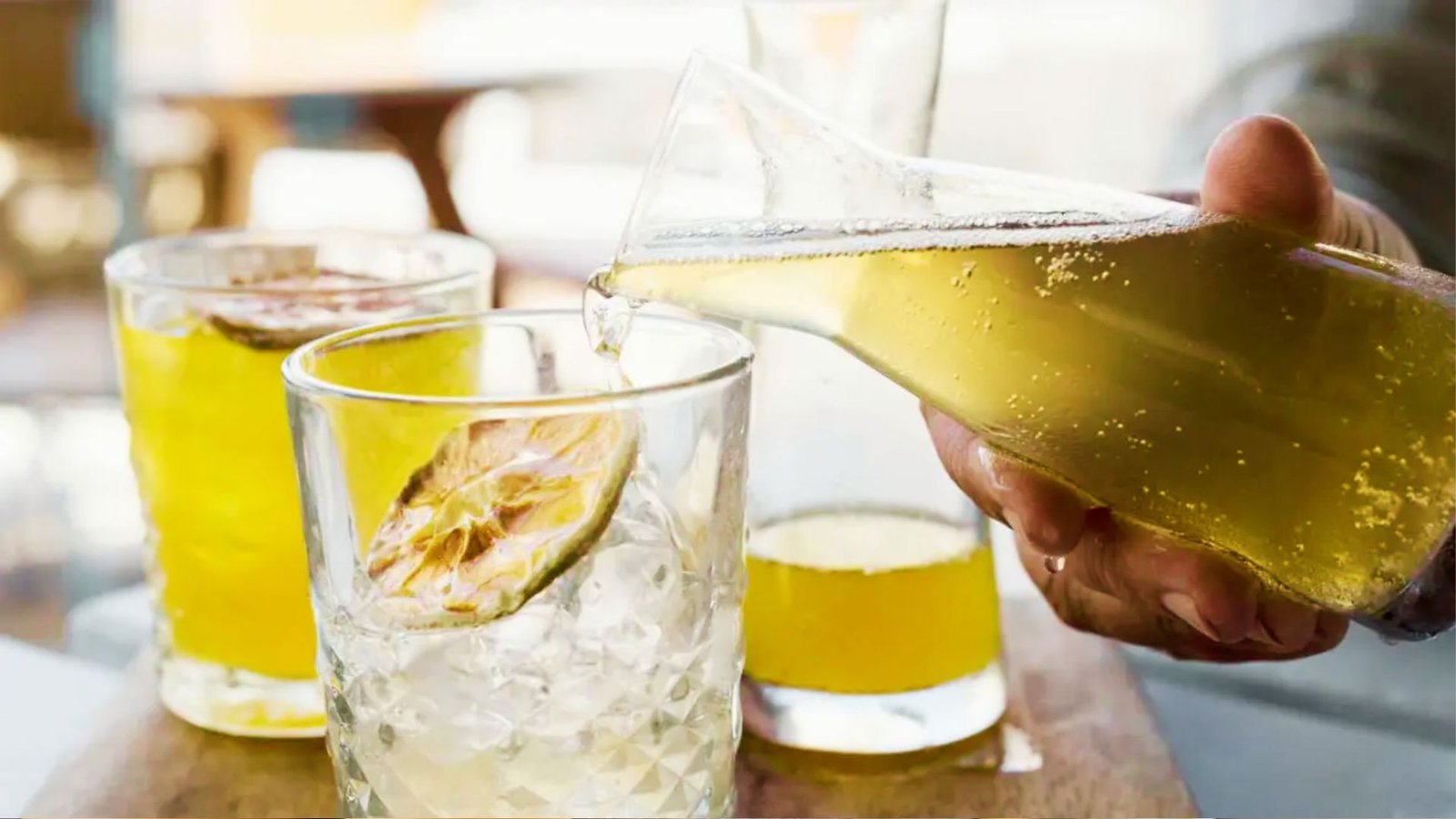 Hand pouring fizzy kombucha from bottle into glass with ice and dried citrus over table.