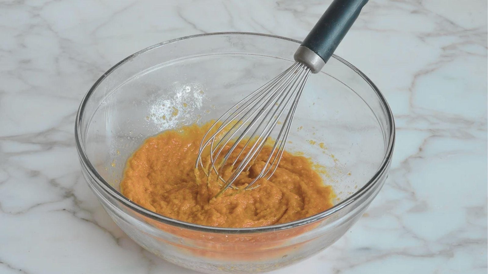 Glass bowl on a marble counter with smooth orange pumpkin puree being mixed with a metal whisk.